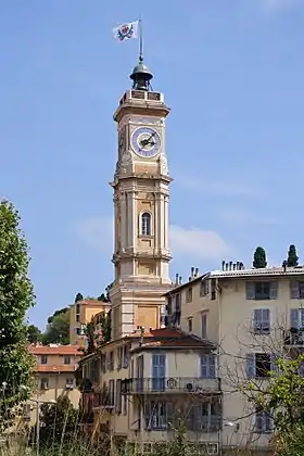 Vue en ville d'une tour avec une horloge. Sur le toit de la tour se trouve une cloche surmontée d'un drapeau.