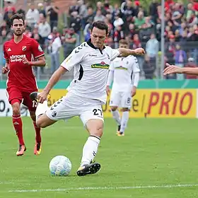 Photographie d’un homme, de pied, en tenue de footballeur, au moment de frapper une balle, la jambe droite en l’air