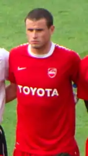 Photographie d'un joueur, portant un maillot rouge et posant pour un cliché. La photo est prise d'assez loin