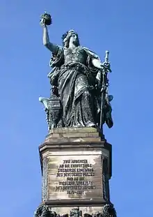 Grande statue de bronze sur un socle en marbre. Elle tient un épée dirigée vers le sol dans sa main gauche et la couronne impériale dans sa main droite qu'elle brandit au ciel. Elle est habillée d'une grande robe aux allures guerrières.