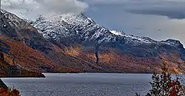 Des montagnes enneigées et recouvertes d'arbres aux couleurs automnales sous un ciel sombre, derrière un lac.