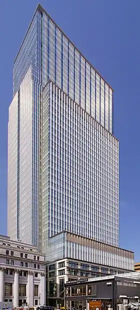 Ground-level view of a rectangular, glass high-rise; adjoining the high-rise is a stone building featuring columns