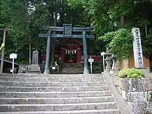 Photo couleur d'un torii en haut d'un escalier en pierre. Des arbres aux feuilles vertes forment l'arrière-plan.