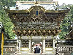 Photo couleur de la porte d'entrée d'un sanctuaire shintō, avec, de part et d'autre de son ouverture, une statue représentant un gardien divin, et, un second étage paré de dorures sous un toit de tuiles. En arrière-plan, une forêt d'arbres au feuillage vert.