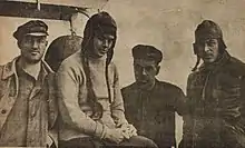 Photographie d'une femme portant un bonnet et des tresses, entre un homme à gauche et deux hommes à droite