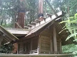 Un bâtiment simple en bois avec une véranda à grumes horizontales sur le faîte du toit et épis de faîtage en forme de fourche. De l'avant, un autre toit s'étend vers la véranda et se relie à un autre bâtiment.