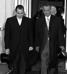 Photographie de deux hommes portant de longues manteaux quittant un bâtiment