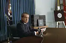 Nixon l'air grave est assis à son bureau devant une pile de classeurs.