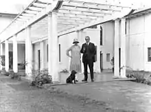 Photographie d'un homme en costume, d'une femme en robe avec un chapeau et un chien noir sous une pergola