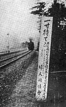 A black-and-white image of a tall signboard with Japanese writing (larger at the top), next to train tracks. There is a train visible in the distance.
