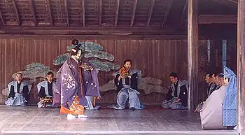 Plafond et mur du fond en bois. Au fond, musiciens accroupis, au premier plan, acteur en kimono chatoyant.