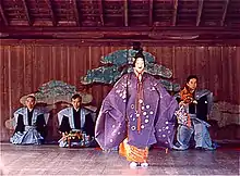 Acteur debout en tenue traditionnelle violette, masque blanc souriant. Trois musiciens visibles en arrière plan.