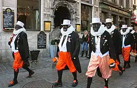 Photographie d'un groupe d'hommes portant un chapeau blanc, une veste de costume noire, un pantalon orange, des bas et des chaussures noirs, le visage peint en noir.