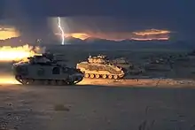 plusieurs véhicule blindé côte à côte au crépuscule, l’un d’eux tire un missile qui éclaire la scène, tandis que les éclairs d’un orage illuminent les montagnes à l’arrière-plan