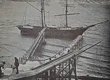 Photographie de deux hommes devant plusieurs blocs de glace sur un ponton en bois au bord de la mer. Plusieurs rampes en bois sur pilotis s'avancent au-dessus de la mer et un navire à deux mats est ancré à l'extrémité de l'un d'eux.