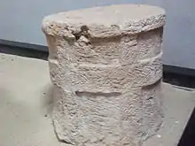 Un cylindre de pierre avec des badndes verticales et une bande horizontale en relief sur le pourtour.