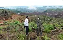 Photographie de deux hommes debout devant un paysage de collines déboisées.