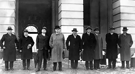 Photo en noir et blanc de neuf hommes en manteau et chapeau debout devant un bâtiment
