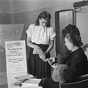Photographie en noir et blanc de deux femmes testant la texture d'un bas, l'une debout et légèrement penchée en avant, l'autre, plus âgée, assise et attentive à la démonstration.