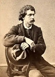 Photographie en noir et blanc d'un homme moustachu, accroupi, portant un large chapeau à sa main gauche.