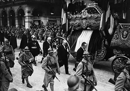 Photo en noir et blanc d'un cortège funéraire accompagnant un imposant véhicule sur lequel est posé un drapeau tricolore