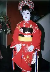 Photographie en couleur d’une danseuse de nihon-buyo en costume traditionnel. Elle porte un kimono rouge. Elle a les cheveux noirs attachés en chignon et le visage maquillé de blanc.