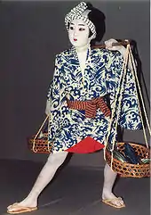 Photographie en couleur d’un danseur de nihon-buyo. Il porte un kimono avec des motifs bleus. Son visage est maquillé de blanc. Il se tient sur une scène en bois et regarde en face de lui.