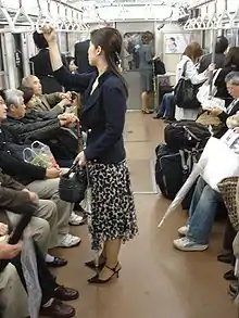 Photo couleur d'une femme debout (premier plan), vue de profil, à l'intérieur d'une rame de métro. Plusieurs personnes sont assises de part et d'autre de la partie centrale de la rame.