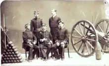 Une photographie représentant un groupe de 5 hommes en uniforme entre une pile d'obus sur la gauche et une pièce d'artillerie à roues sur la droite
