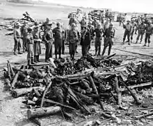 Groupe d'hommes en uniforme devant les restes d'un bûcher où se trouvent les restes calcinés de corps humains.