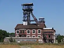 Photographie en couleur représentant, au premier plan, un long entrepôt de brique à un niveau, au deuxième plan un bâtiment cathédrale surmonté d'un chevalement de mine, et au troisième plan le sommet d'un second chevalement