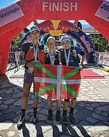 Trois femme tenant un drapeau basque devant une arche d'arrivée rouge.