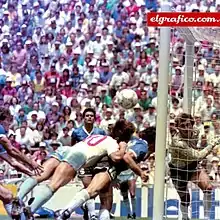 Photographie en couleurs. Un joueur anglais et un joueur argentin se jettent presque à l'horizontale vers le ballon qui file vers les buts laissés vides par le gardien argentin, en retard.