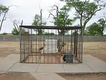 Trois tombes avec une seule pierre tombale. La tombe de Billy the Kid, à droite, possède en plus une petite stèle pointue, renforcée de métal. L'ensemble est enfermé dans une grande cage en métal et cerné de dalles de béton. Le sol du reste du cimetière est nu, apparemment vide, avec un mur à l'arrière plan.