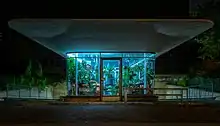 Un minuscule bâtiment entièrement vitré surmonté d'un grand auvent plat remplit de plantes. Il fait nuit, et toute la lumière diffuse depuis l'intérieur de ces plantes par un éclairage artificiel.