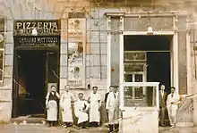 Photo ancienne et jaunie, on distingue au moins huit hommes posant côte à côte devant le restaurant. A gauche de la photo, on peut lire au-dessus de l'entrée Pizzeria internazionale Gennaro Mattozzi.