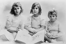 Photographie en noir et blanc de trois petites filles vêtues de blanc et assises devant un livre ouvert.