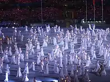 Photographie d'une scène d'un spectacle, de nombreux danseurs au centre en combinaison blanche.