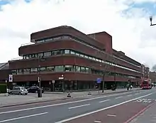 Extérieur d'un grand bâtiment de trois étages en briques rouges et en verre à côté de la route