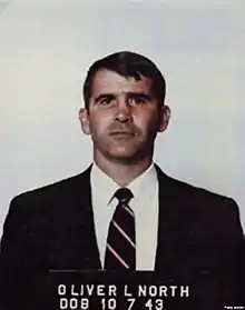 Photographie en couleurs et en buste d'un homme derrière une plaque de détention et portant un costume
