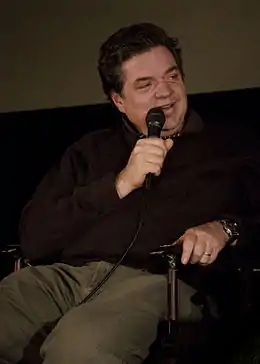 Photographie d'un homme d'une cinquantaine d'années assis dans un fauteuil. Il tient un microphone dans sa main droite et porte une montre à sa main gauche.