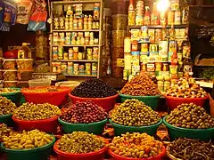 photo en couleurs, au premier plan des bassines en plastique remplies d'olives et de piments, au fond des étagères portent des boites de conserve et des bouteilles d'huile