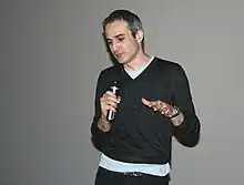 alt=Homme de 53 ans, mince, portant un pull gris sur un T-shirt clair près du corps, courts cheveux gris, qui parle dans un micro