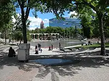 Photo de la Place olympique. On voit des arbres, un bassin plein d'eau et, en arrière-plan, des bâtiments