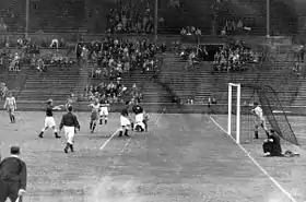 Photographie en noir et blanc d'une scène d'un match de football avec le but à droite.