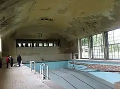 Une piscine abandonnée au toit incurvé. Des grandes fenêtres font la longueur de la piscine, et des plus petites sont au fond. Trois personnes non identifiables se tiennent debout sur le rebord de la piscine.