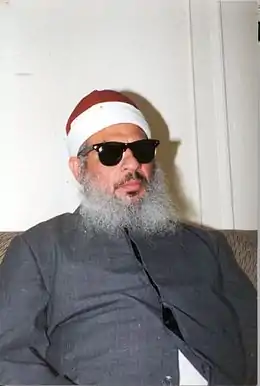 Photographie d'un homme assis sur un canapé avec une barbe blanche, portant des lunettes de soleil et un bonnet.