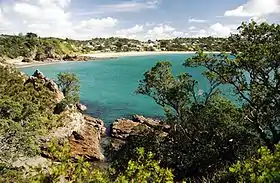 Une baie : au premier plan, au bord de l'eau, des rochers et de la végétation ; au fond une plage et des collines avec des habitations.