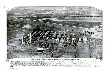 Photo en noir et blanc qui montre un cratère à l'avant-plan. À l'arrière apparaissent des installations détruites puis des immeubes debout.
