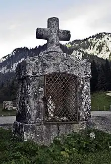 Oratoire en pierre de forme cubique avec une niche contenant une statue de vierge dont l'ouverture est protégée par une grille, le tout surmonté d'une croix.
